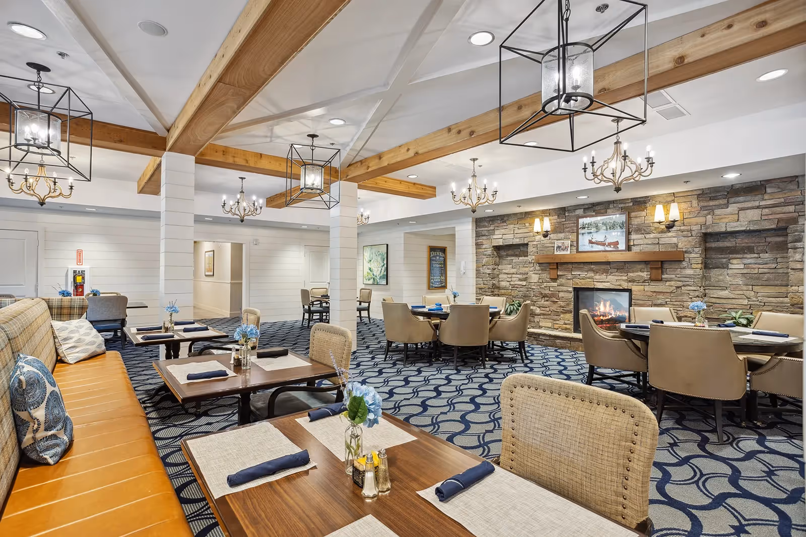 A spacious dining room with multiple tables set with placemats, napkins, and small flower vases. The room features a patterned carpet, a stone fireplace with a wooden mantel, and several chandeliers and pendant lights hanging from a ceiling with exposed wooden beams. There are cushioned chairs and a long bench with pillows along one side.