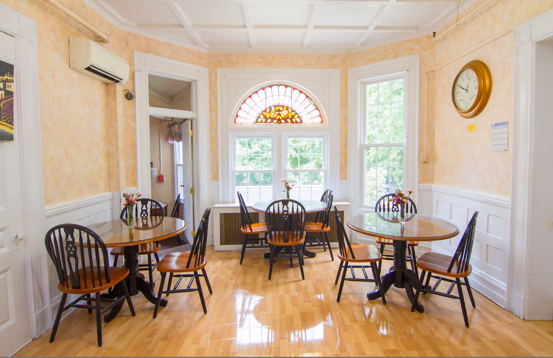Bright dining room with three round wooden tables and chairs on a polished hardwood floor beneath a decorative stained-glass fanlight window.