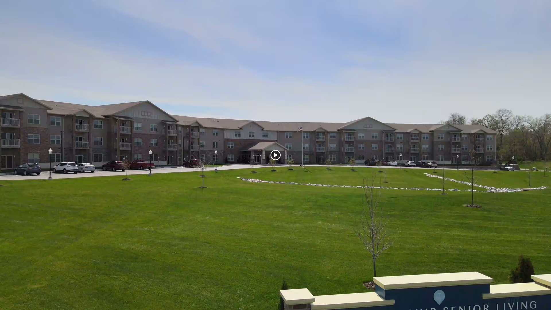 Wide exterior view of a large three-story senior living facility building with multiple balconies and a parking area in front. The foreground features a well-maintained green lawn with young trees and a stone-lined pathway. The sky is clear with some light clouds.