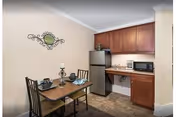 Small dining area with a wooden table set for two, featuring two chairs, a decorative candle holder, and tableware. Adjacent to the dining area is a compact kitchen with wooden cabinets, a stainless steel refrigerator, microwave, and coffee maker. The walls are light-colored with a decorative wall piece above the table.