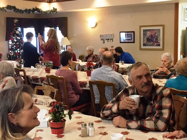 A group of elderly people and a few younger adults gathered in a decorated common room with tables covered in festive tablecloths and small holiday-themed centerpieces. A decorated Christmas tree is visible in the background near a window. People are sitting and conversing, and one man in the foreground is holding a cup.