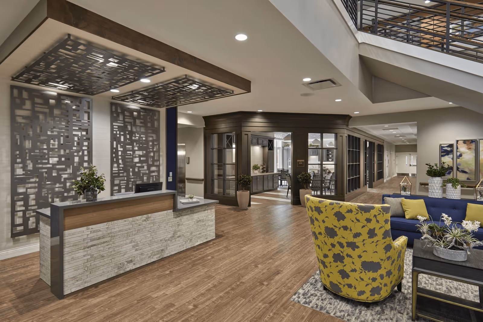 A modern senior living facility interior featuring a reception desk with decorative panels behind it, a seating area with a yellow floral armchair and a blue sofa with green pillows, wooden flooring, and a hallway leading to other rooms. The space is well-lit with recessed ceiling lights and has a contemporary design with plants and artwork on the walls.