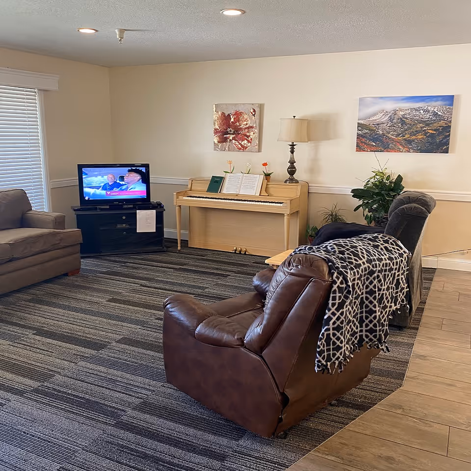 Bright living room in a senior facility with a piano, TV, seating, and artwork.