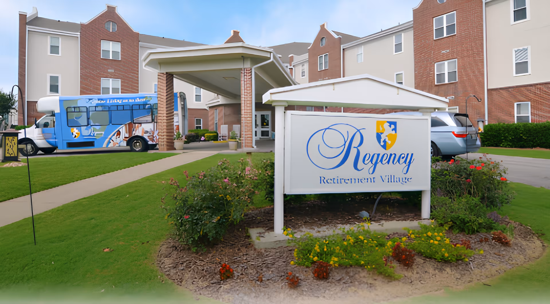 Exterior view of Regency Retirement Village of Birmingham showing the main entrance with a covered drop-off area, a blue shuttle bus parked on the left, a silver vehicle on the right, and a landscaped garden with flowers in front of the facility sign.