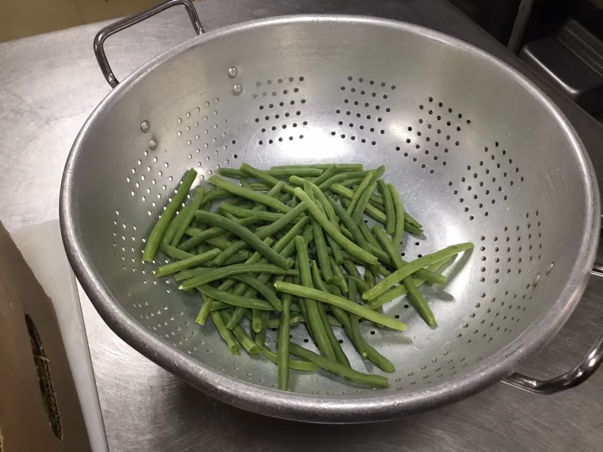 Metal colander holding trimmed green beans on a stainless steel kitchen counter.