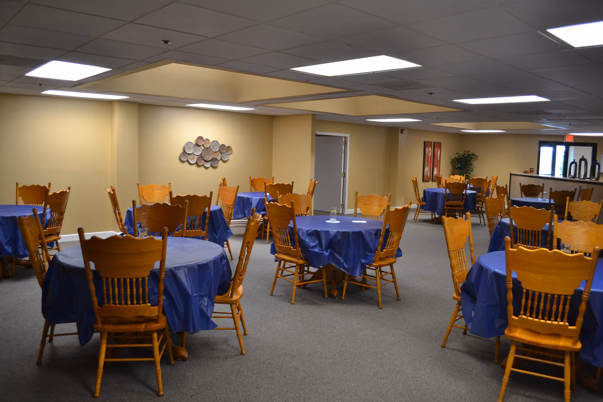 Interior view of a dining room with multiple round tables covered in blue tablecloths, each surrounded by wooden chairs. The room has beige walls, ceiling lights, and some wall decorations including a metal wall art piece and framed pictures. There is a counter area visible in the background near an exit door.