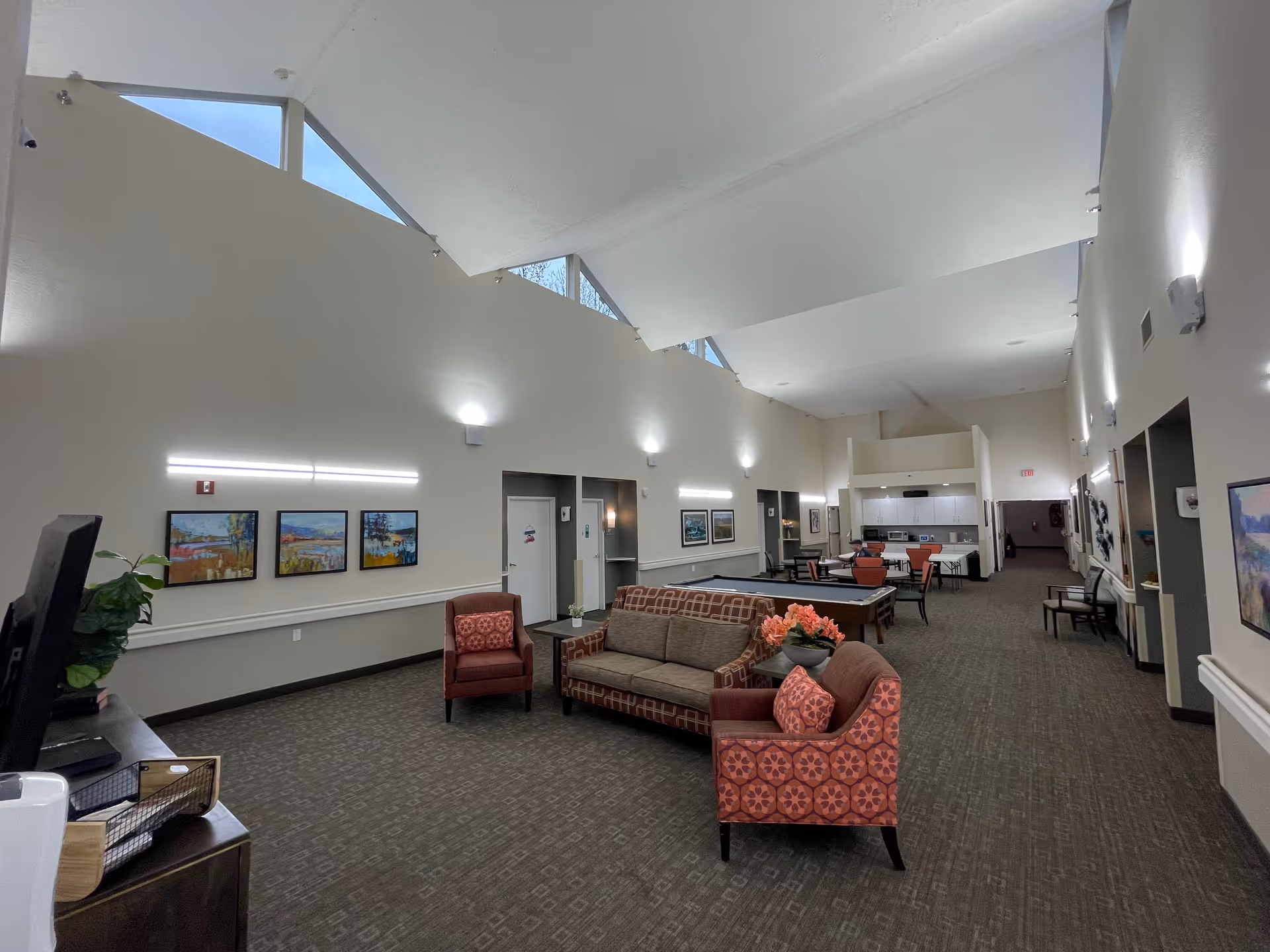 A spacious senior living common area with high ceilings and triangular windows near the ceiling. The room features a seating area with a sofa and two patterned armchairs, a pool table, and a dining table with chairs. The walls are decorated with framed artwork and illuminated by modern wall lights. There is a kitchenette area in the background.