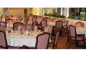A dining room with multiple round tables covered with white tablecloths, set with glassware, plates, and napkins. The chairs around the tables have cushioned backs and seats. The room has large windows with curtains and some greenery along the walls.