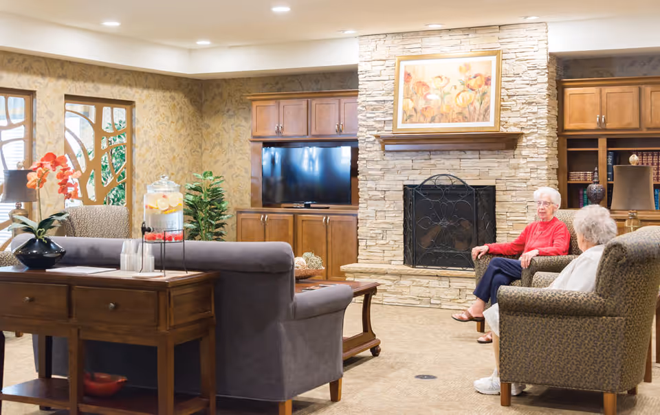 Common room of a senior living facility with sofas and armchairs around a stone fireplace and TV, two elderly women seated and conversing.