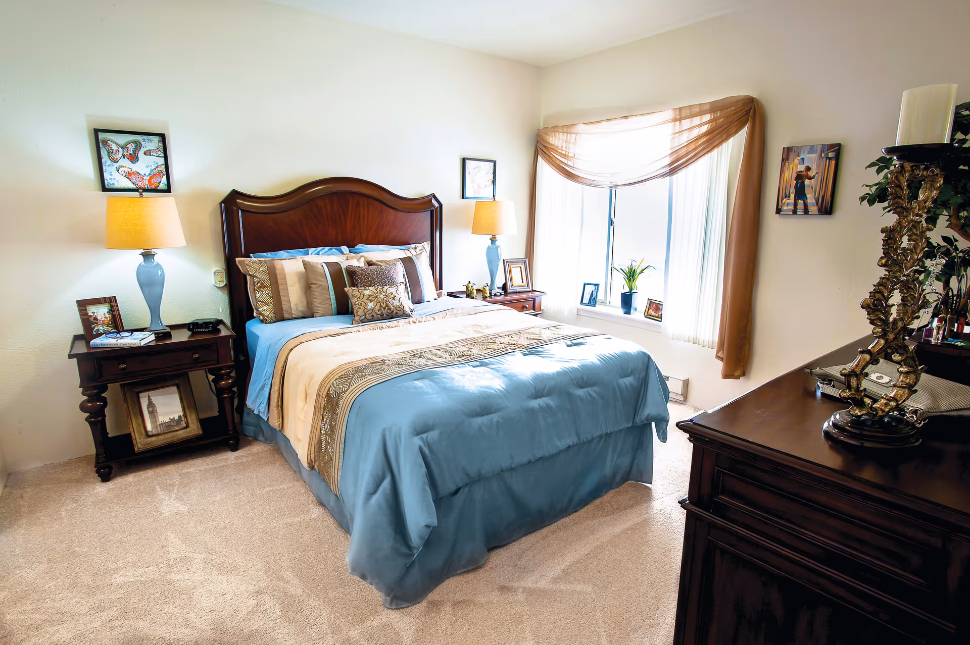 A cozy bedroom with a large wooden bed featuring blue and beige bedding. There are two wooden nightstands on either side of the bed, each with a lamp and framed pictures. A window with sheer curtains and a brown valance lets in natural light. A dark wooden dresser with decorative items is visible on the right side of the room.
