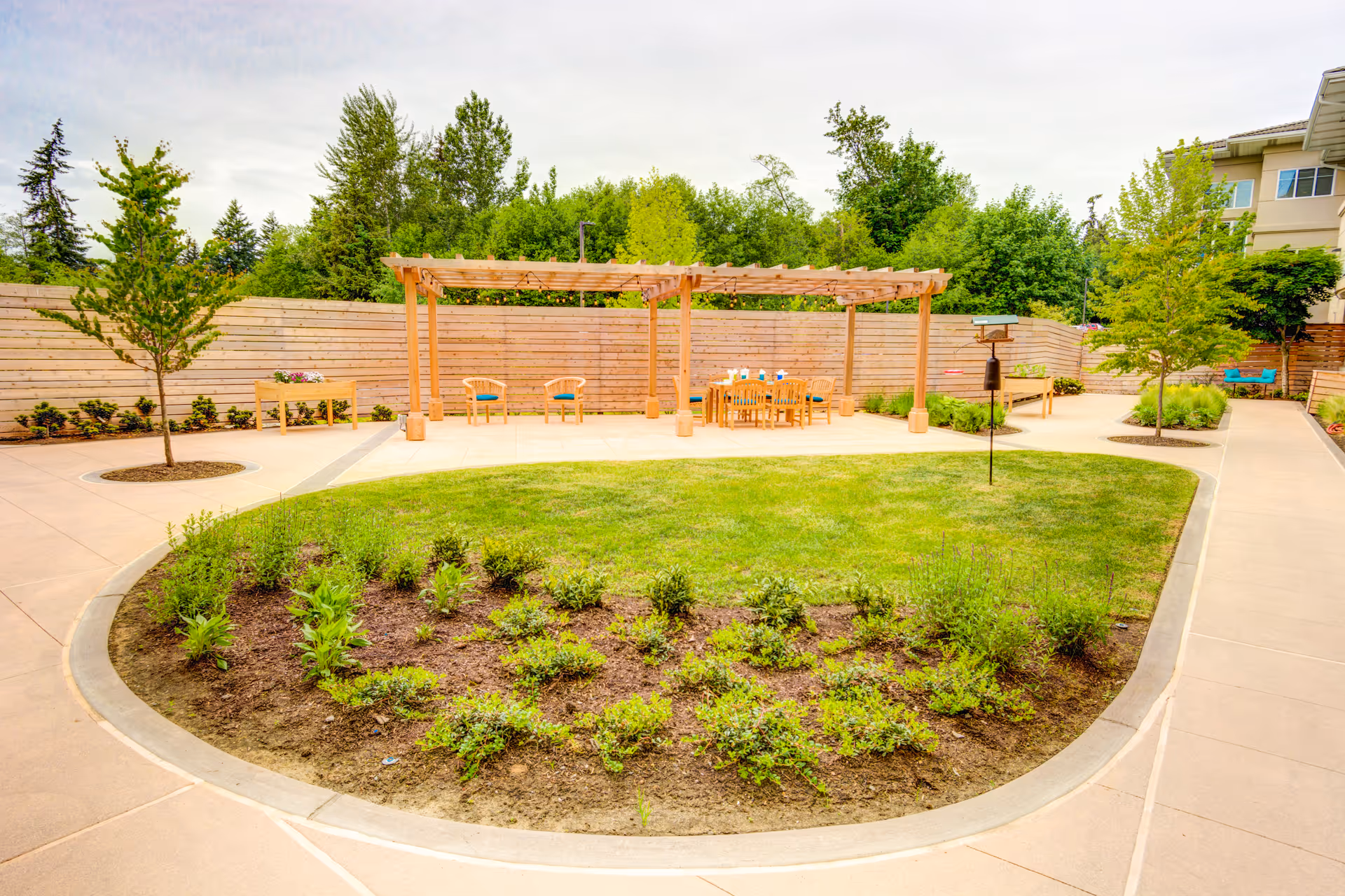Outdoor garden area at Harbour Pointe Senior Living featuring a curved flower bed with small plants, a grassy lawn, paved walkways, young trees, and a wooden pergola with chairs and a table underneath. A wooden fence and dense green trees are visible in the background.
