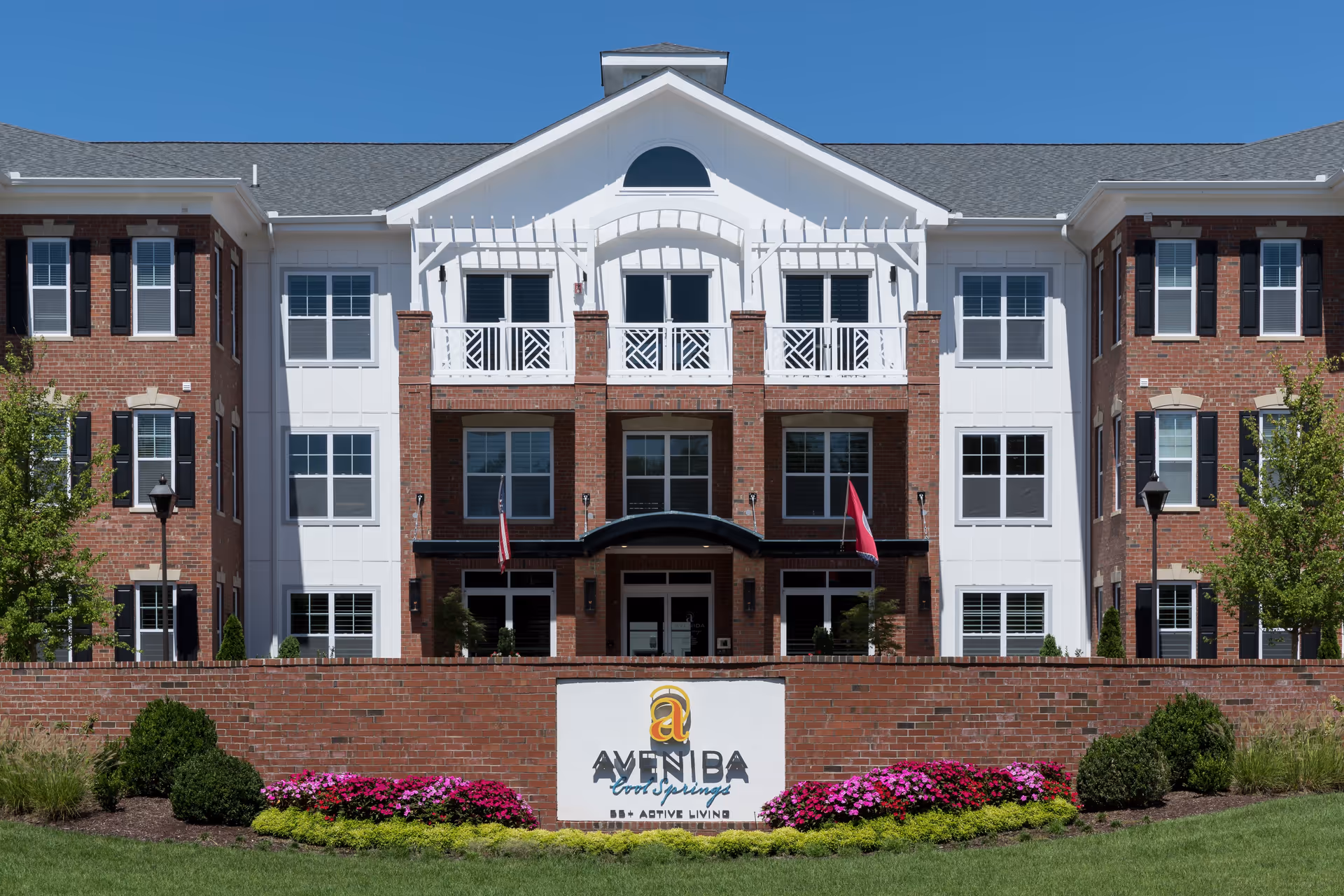 Front exterior view of Avenida Cool Springs building with a brick facade, white accents, multiple windows, and a sign in front surrounded by landscaping and flowers under a clear blue sky.