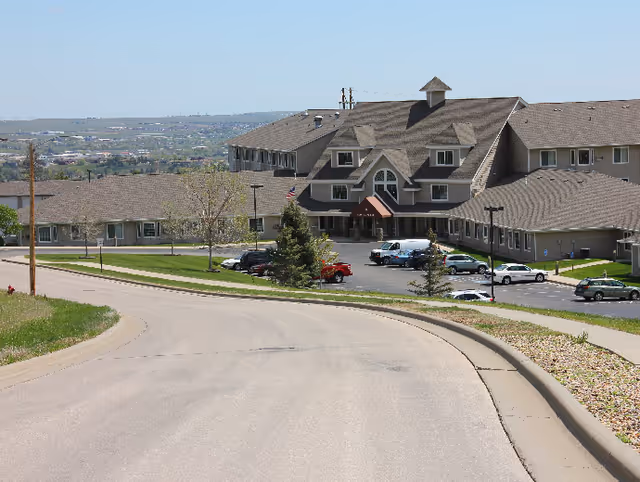 View of the Good Samaritan Society - Echo Ridge - The Manor building from the road, showing a large multi-story facility with a peaked roof, several parked cars in front, and a clear sky in the background.