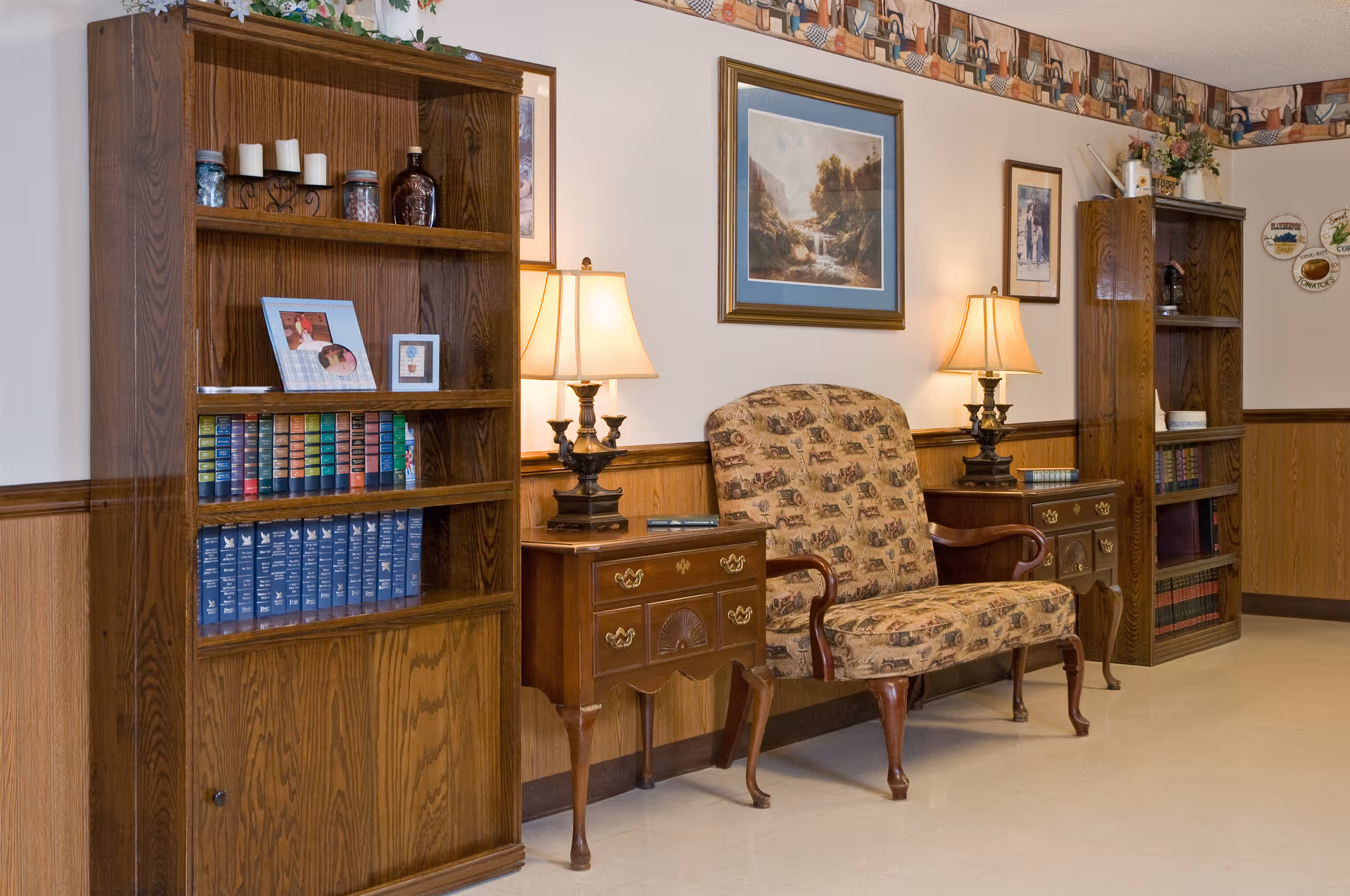 A cozy interior room with two wooden bookshelves filled with books and decorative items, a patterned upholstered loveseat between two wooden side tables with lamps, and framed artwork on the wall above the loveseat.