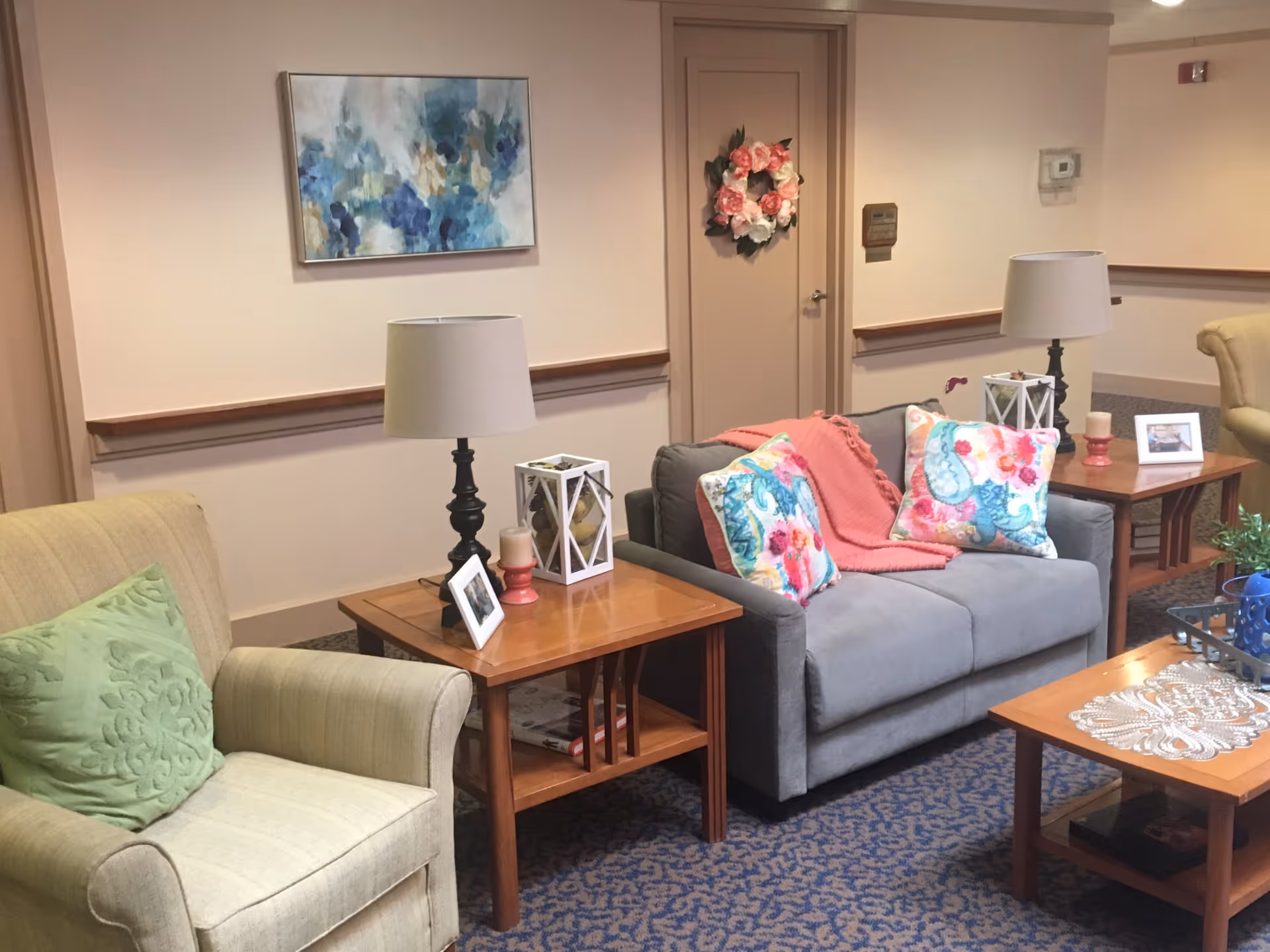 A cozy common lounge with upholstered armchairs, a gray loveseat adorned with colorful pillows and a throw, wooden side tables with lamps, and wall art.