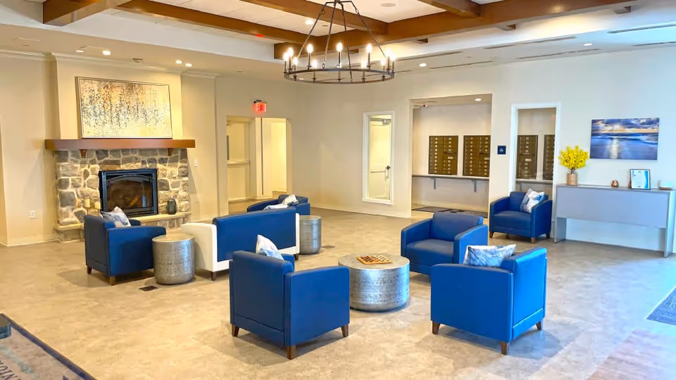 A spacious senior living facility common area with blue and white armchairs arranged around two round metallic coffee tables. There is a stone fireplace with a painting above it on the left wall. The room has beige walls, a light-colored floor, and a ceiling with wooden beams and a circular chandelier. On the right side, there is a console table with a vase of yellow flowers and a framed picture above it. Mailboxes are visible in an alcove in the back wall.