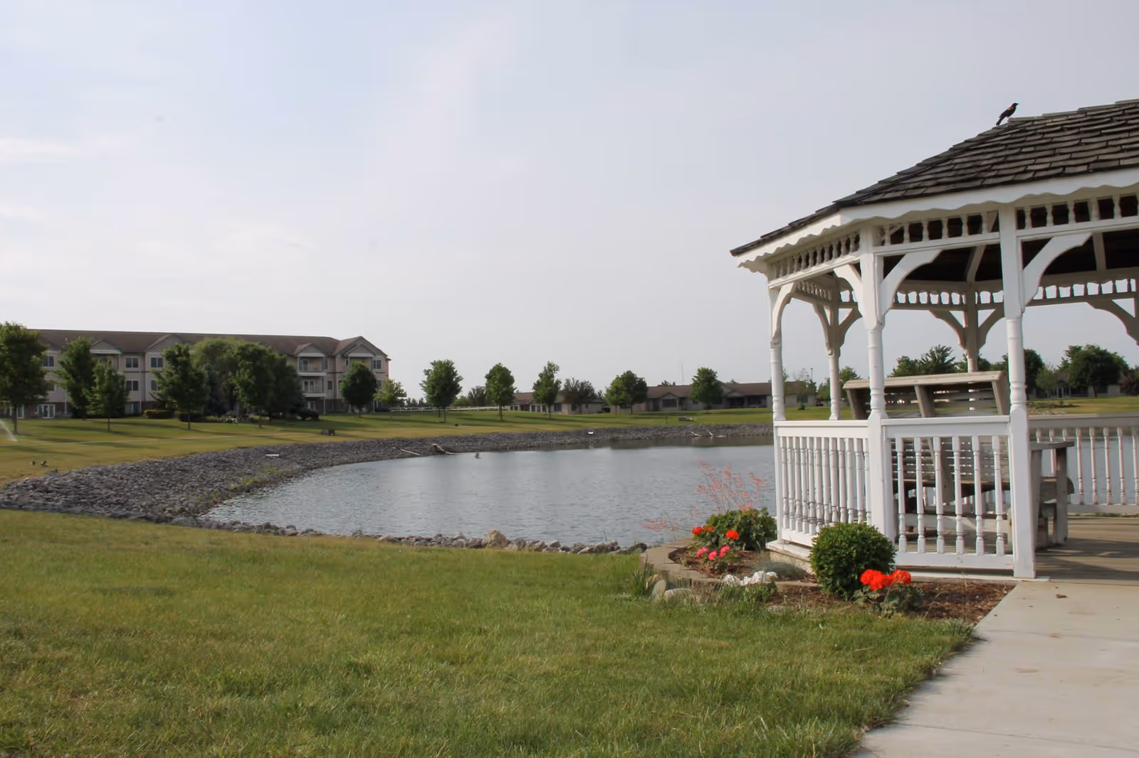 A peaceful outdoor scene at Greencroft Goshen featuring a white wooden gazebo with a shingled roof on the right side, surrounded by green grass and flower beds with red and pink flowers. In the background, there is a small pond bordered by rocks, trees, and a multi-story residential building under a partly cloudy sky.