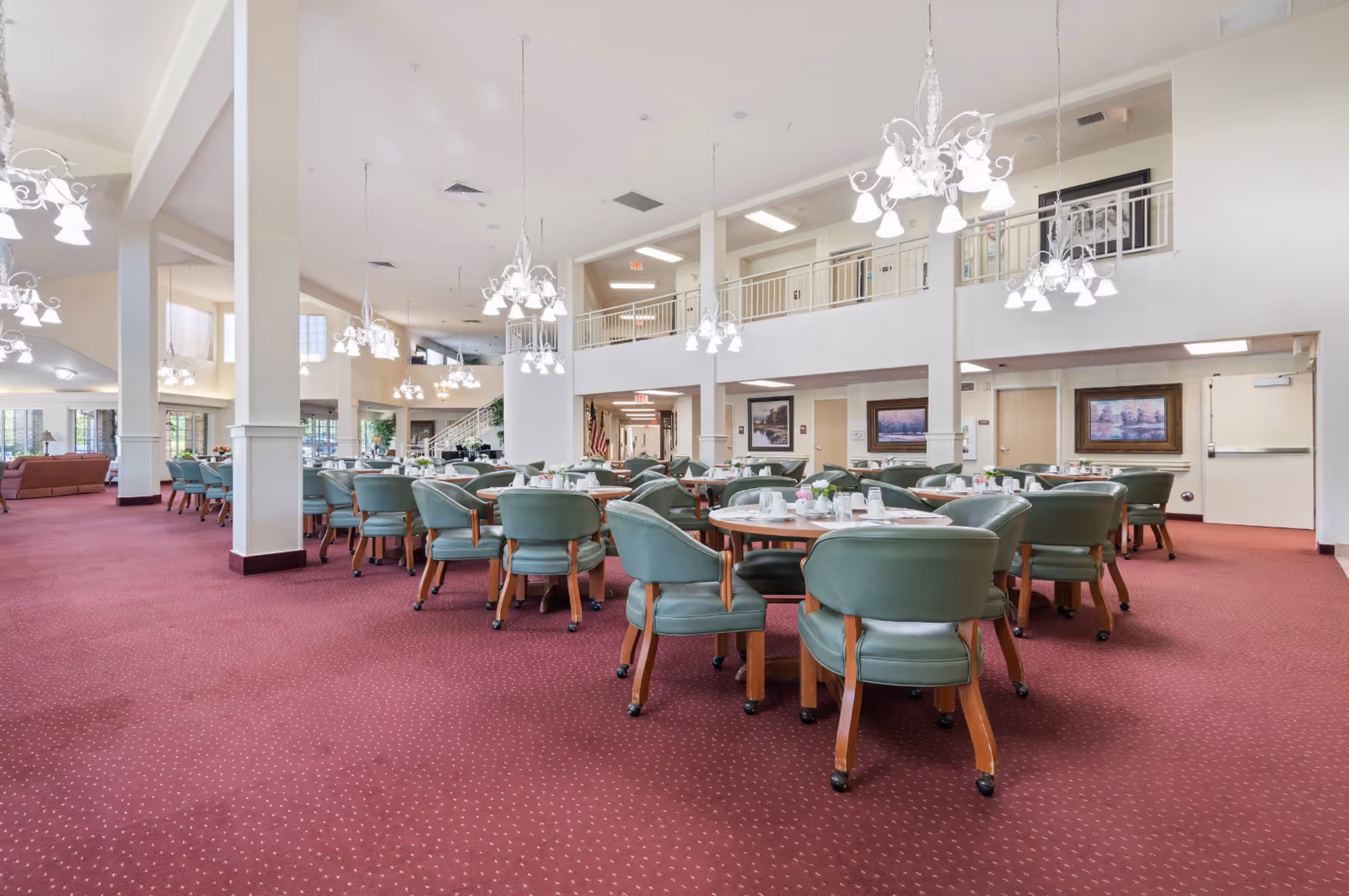 Spacious two-story dining room with round tables set for meals, green chairs, chandeliers, and a red carpeted floor.