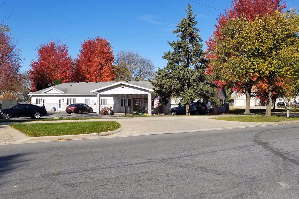 Exterior view of a single-story white building with a gray roof, surrounded by trees with autumn foliage in red, orange, and green. Several cars are parked near the building, and an American flag is visible near the entrance. The scene is set on a clear day with a blue sky.