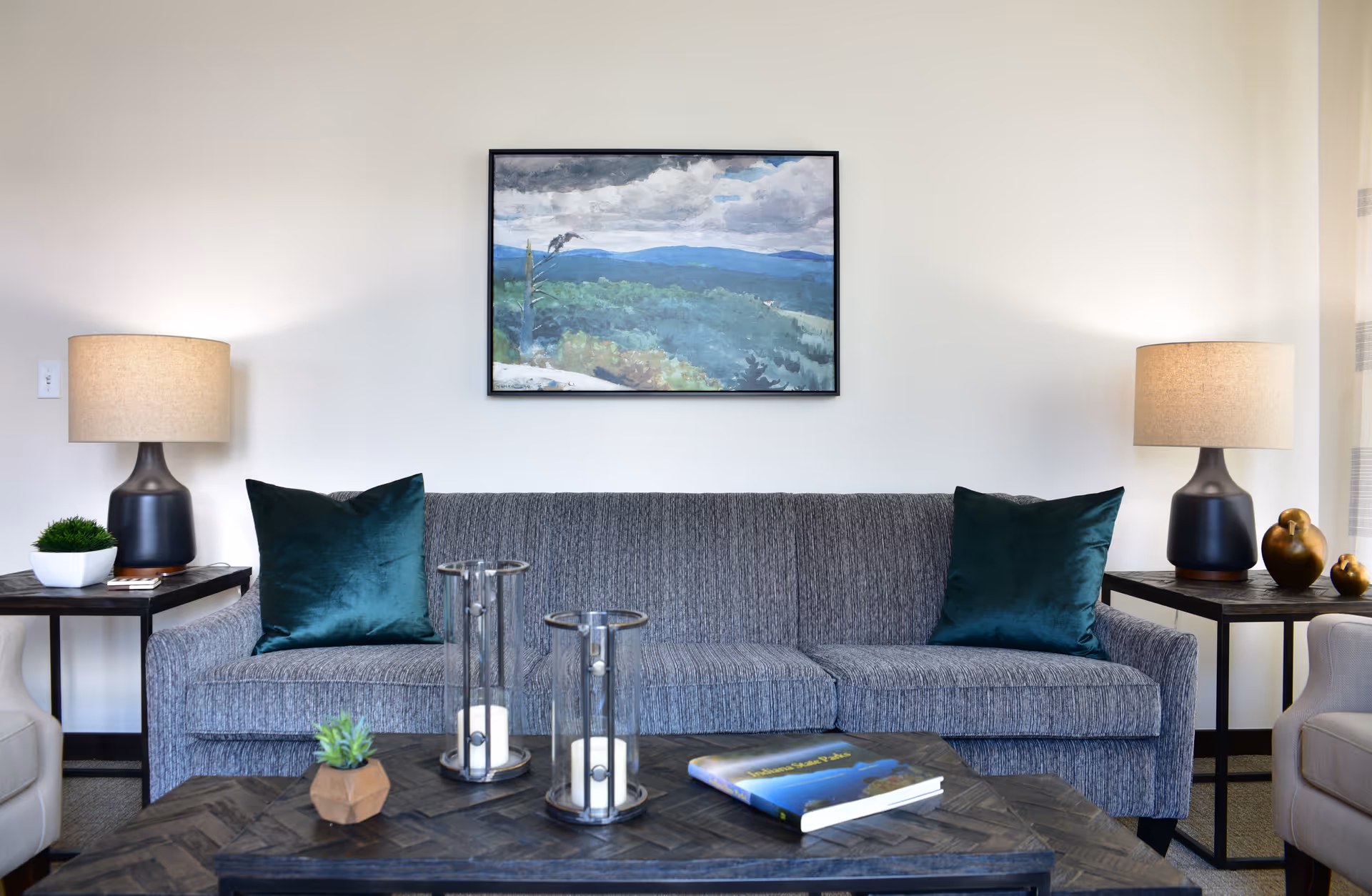 Gray sofa with teal throw pillows in a living room, flanked by side tables and lamps, with a coffee table and a landscape painting above.