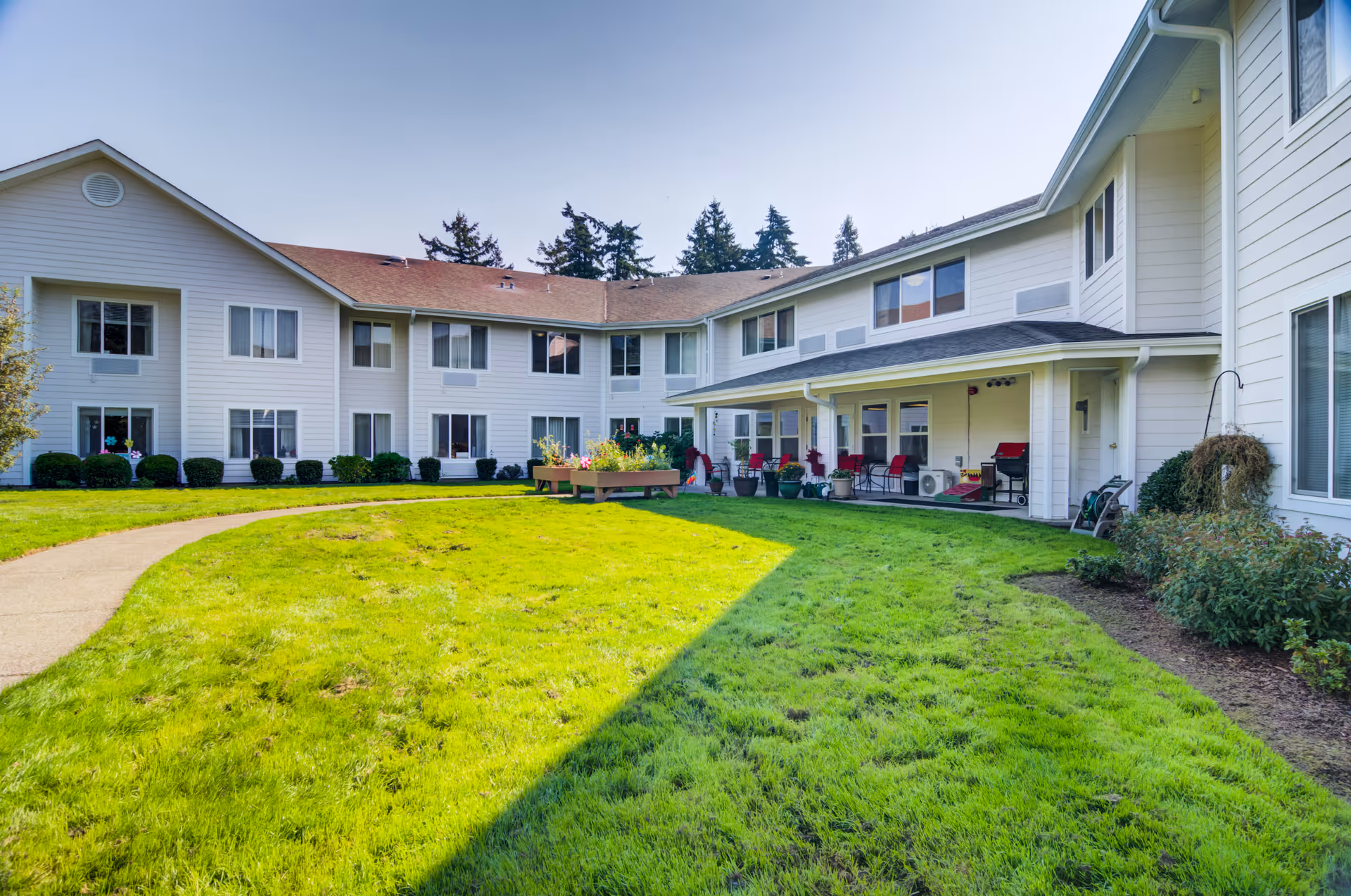 Outdoor courtyard area of a senior living facility with a well-maintained green lawn, a curved concrete pathway, and a two-story white building surrounding the courtyard. There are bushes along the building and a raised garden bed with flowers in the center. The sky is clear and blue.