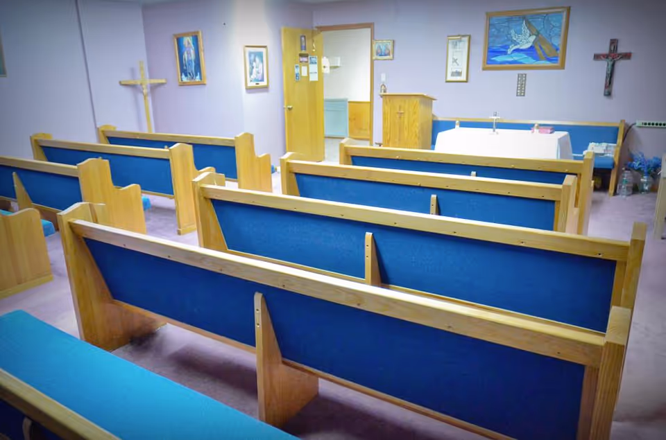 Interior view of a small chapel or prayer room with wooden pews featuring blue cushions, religious paintings and crosses on the walls, a wooden podium, and a table covered with a white cloth at the front.