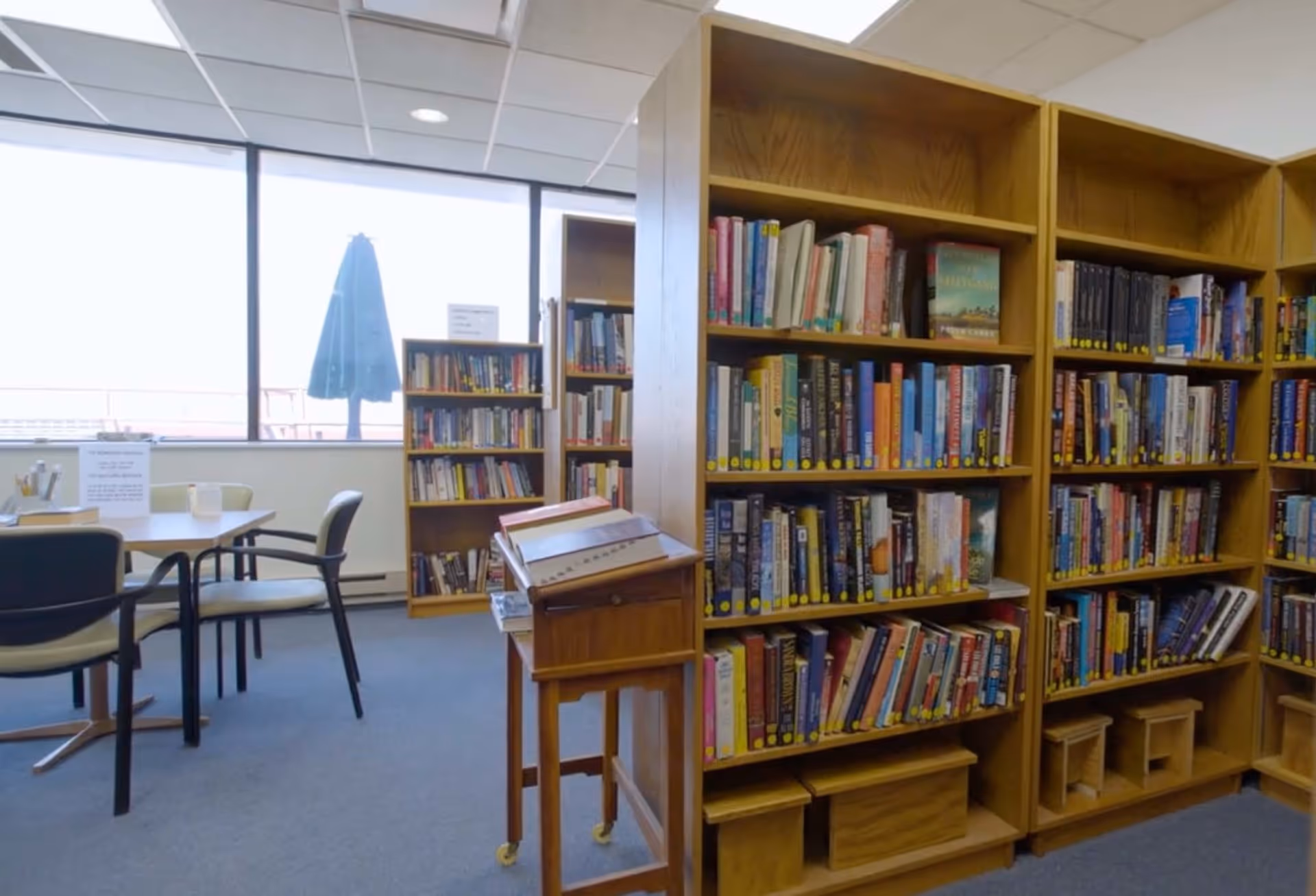 Interior view of a library or reading room with wooden bookshelves filled with books, a table with chairs near large windows, and a wooden stand with a display on it.