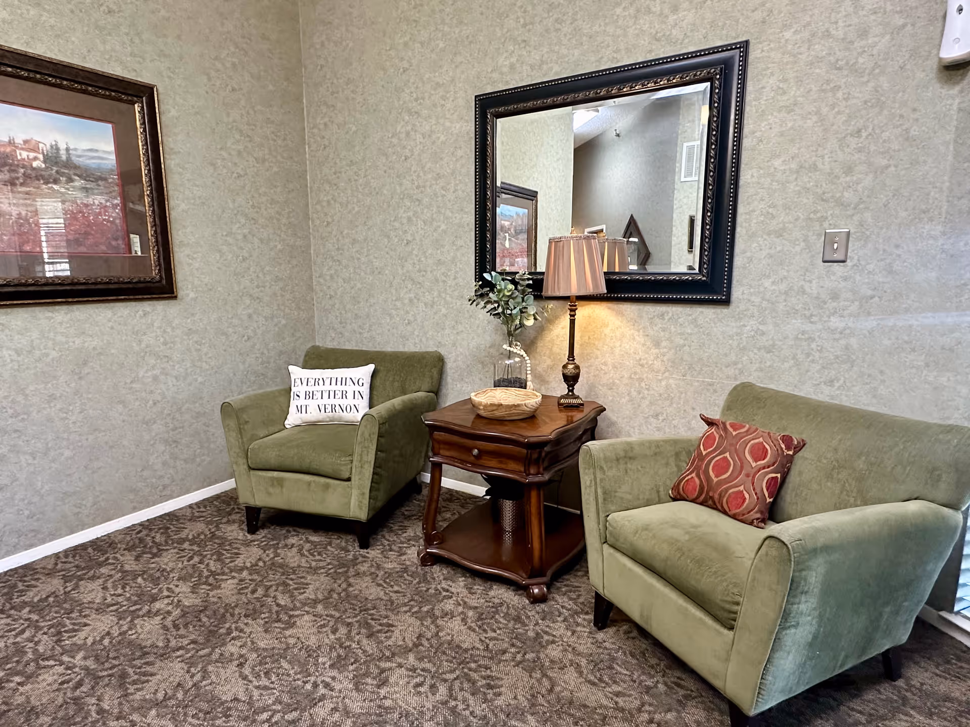Cozy seating area with two green armchairs flanking a wooden side table topped by a lamp and decor beneath a large wall mirror.