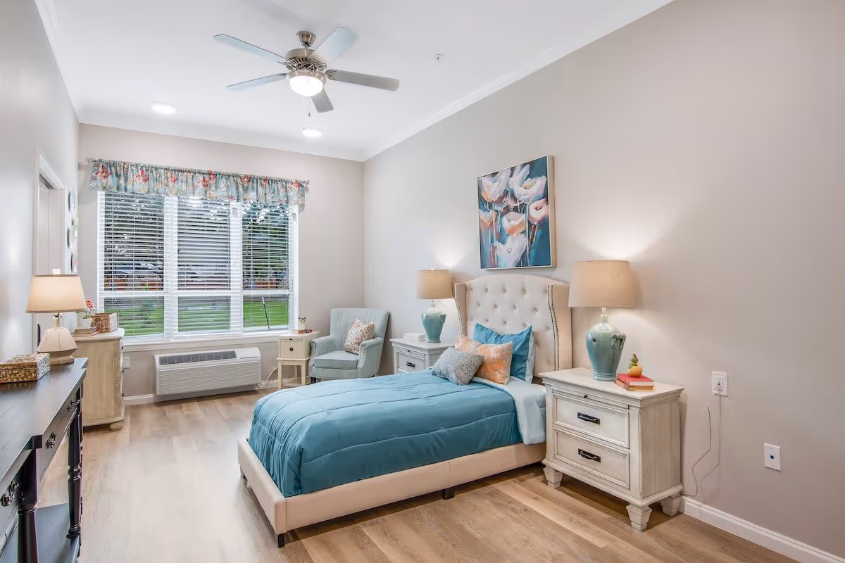 Bright furnished bedroom with a twin bed, nightstands and lamps, an armchair by a large window and a ceiling fan.