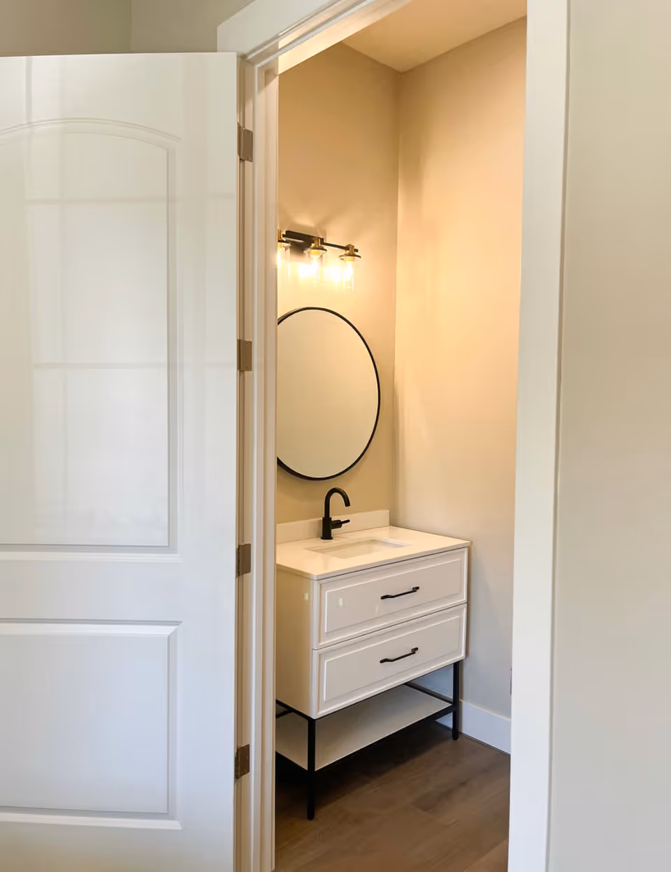 View through a partially open white door into a small bathroom area featuring a white vanity with two drawers, a black faucet, a round mirror with a black frame, and a light fixture with three exposed bulbs above the mirror. The walls are painted beige and the floor is wooden.