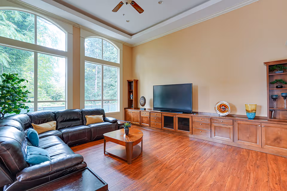 A spacious living room with large windows letting in natural light, a dark brown leather sectional sofa with colorful cushions, a wooden coffee table with a small plant, and a long wooden entertainment unit with a flat-screen TV and decorative items.