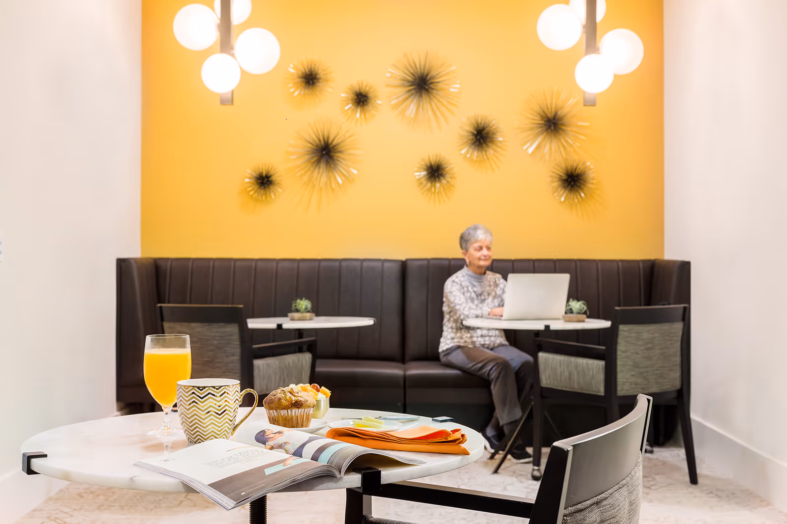 A senior woman works on a laptop at a small table in a bright common dining area, with a foreground table holding a glass of orange juice, a mug, and a muffin.