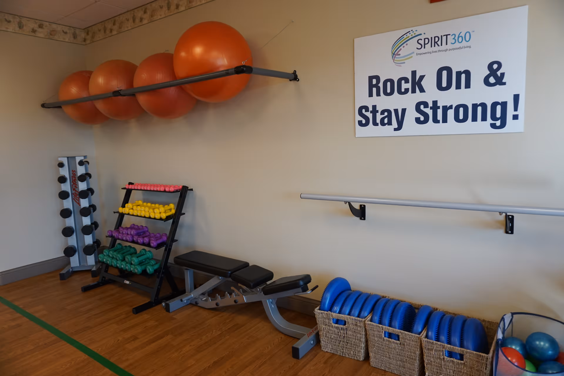Exercise room with stability balls mounted on a rack, colorful dumbbells on a stand, an adjustable bench, baskets of balance discs, and a wall sign that reads 'Rock On & Stay Strong!'