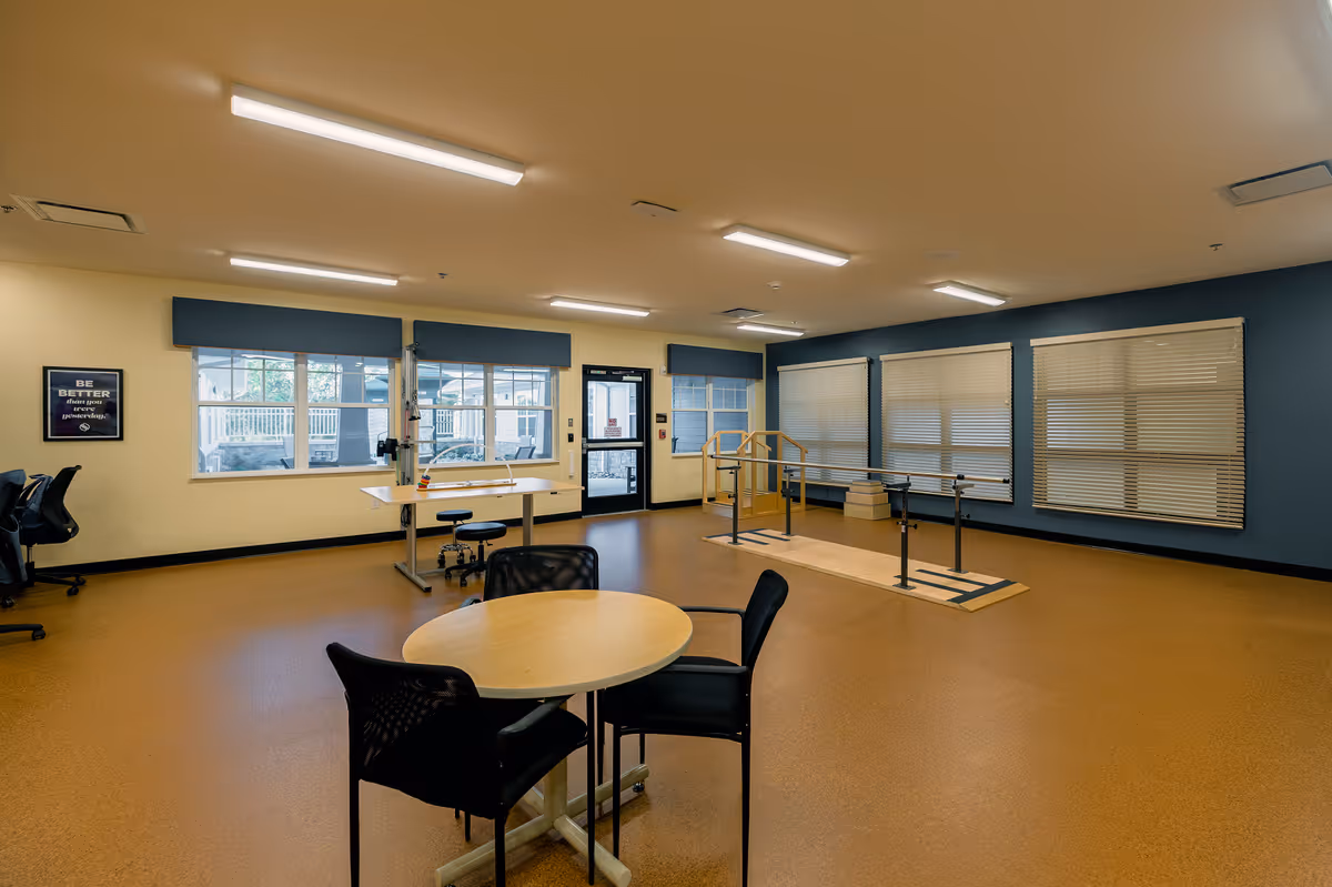 A spacious room with a round table and chairs in the foreground, a desk with stools near the windows, and a set of parallel bars with steps for physical therapy or rehabilitation. The room has large windows with blinds, beige walls with a blue accent wall, and bright overhead lighting.