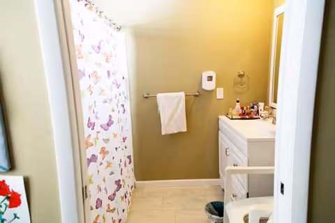 Small senior living bathroom with butterfly-pattern shower curtain, white vanity, towels, and mirror.
