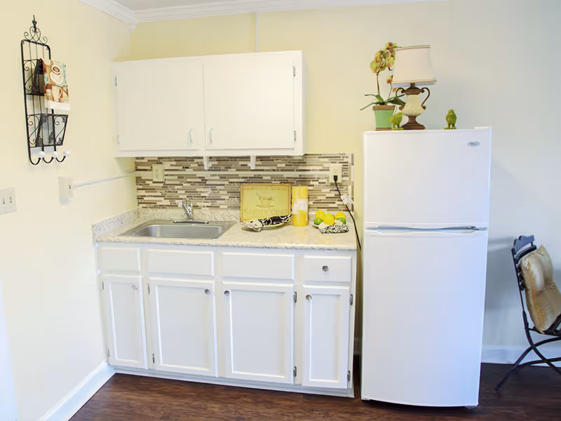 Small kitchen area with white cabinets, a stainless steel sink, a white refrigerator, and a tiled backsplash. There is a decorative wall rack with towels on the left wall and a chair with cushions on the right side. The countertop has a small lamp, a plant, a decorative sign, and some fruit.