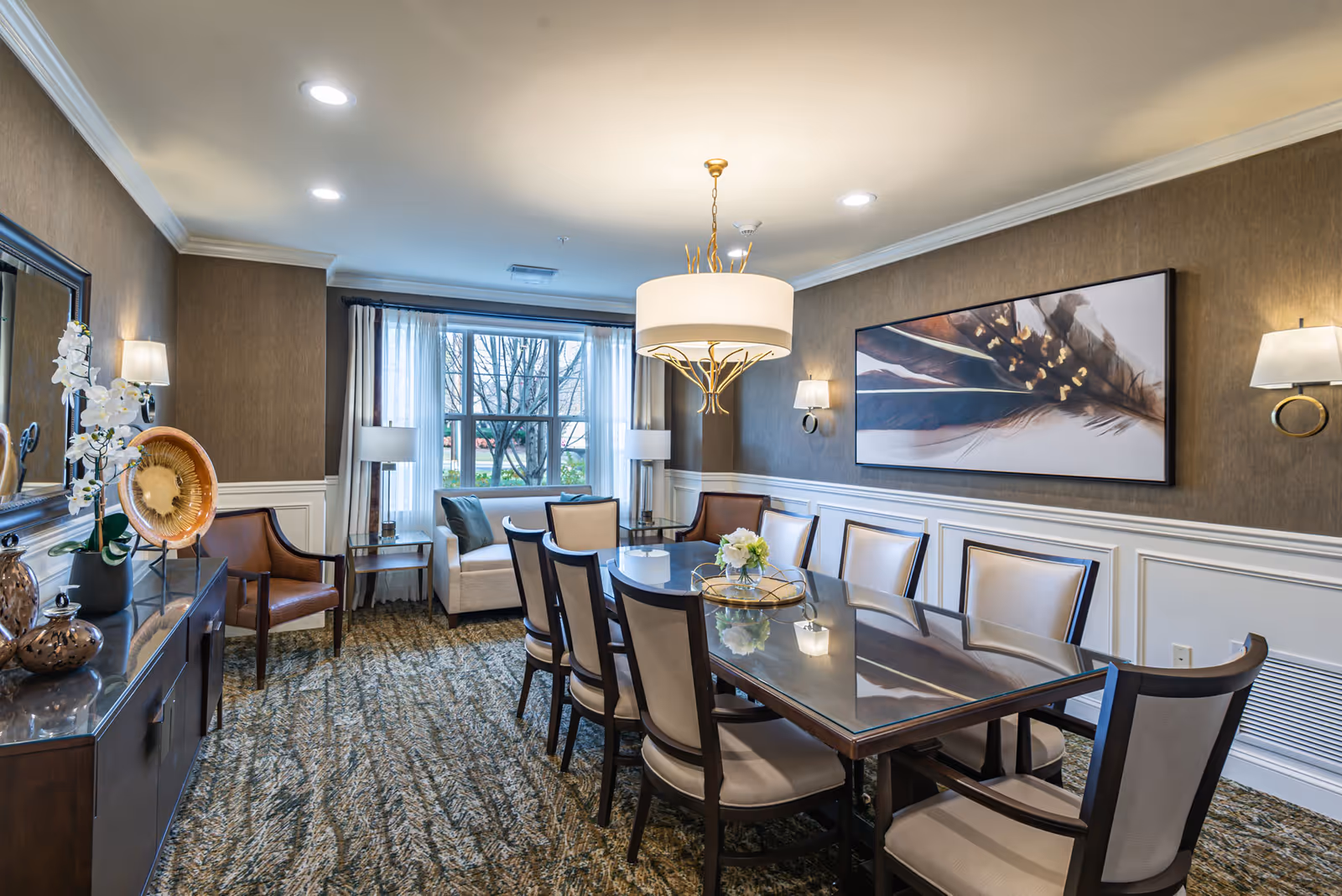 A well-lit dining room with a long glass-top table surrounded by eight cushioned chairs. The room features a large window with white curtains, two armchairs and a small sofa near the window, a chandelier hanging above the table, wall sconces, a large framed artwork of feathers on the wall, and decorative items on a sideboard with a mirror above it.