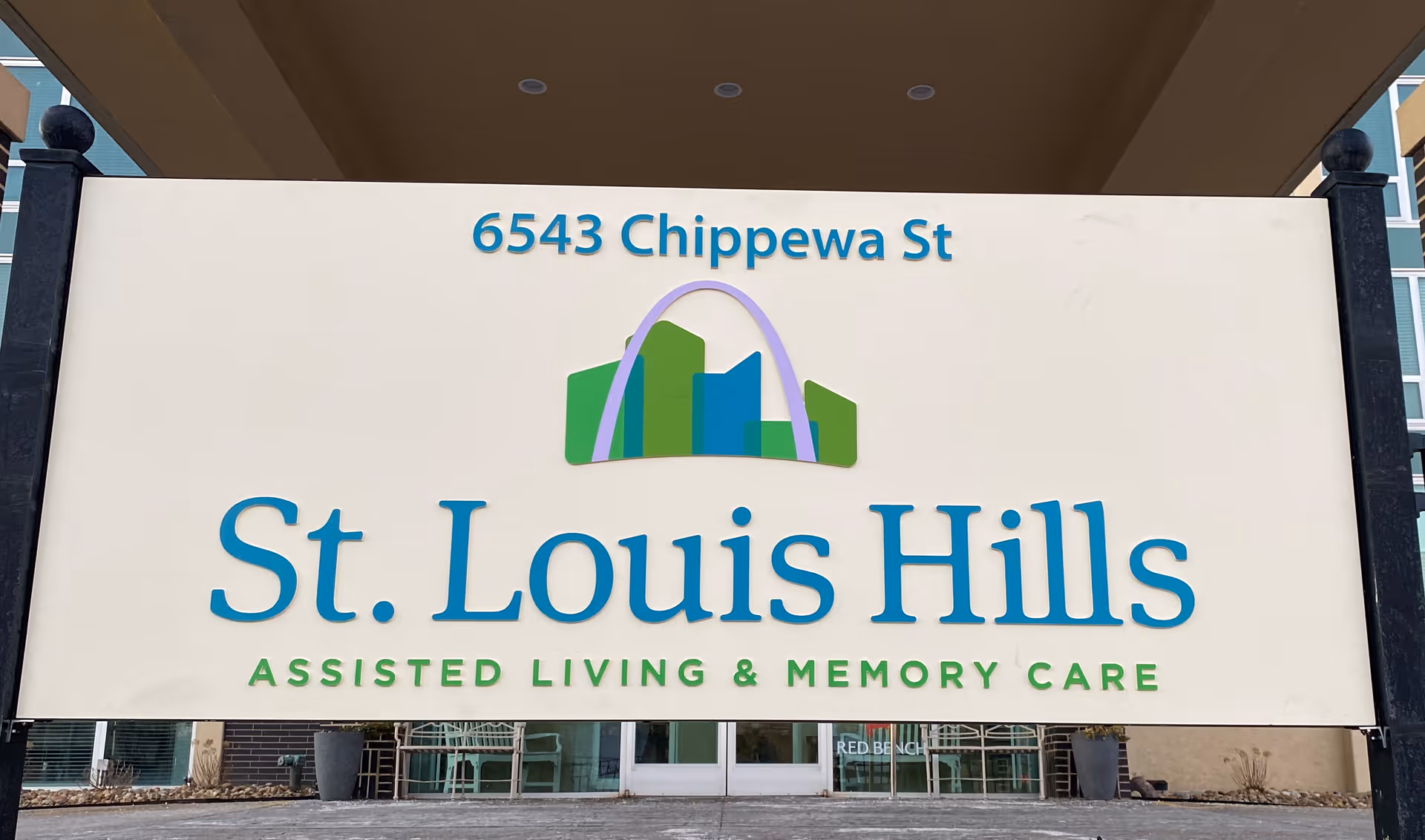 A large outdoor sign for St. Louis Hills Assisted Living & Memory Care located at 6543 Chippewa St. The sign features a stylized graphic of buildings with an arch above them.