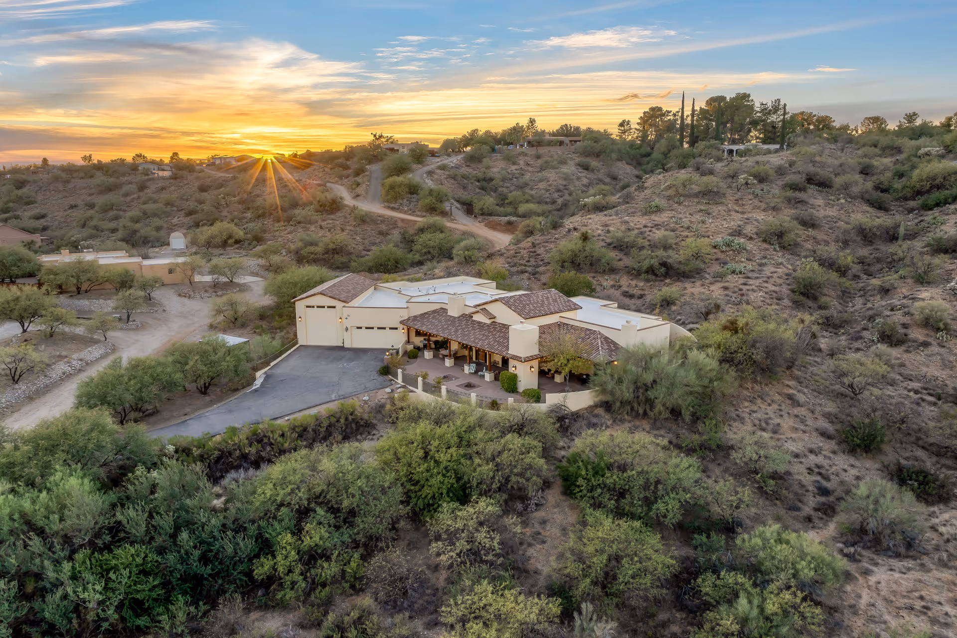 Aerial view of a single-story building with a tiled roof surrounded by desert landscape and hills at sunset. The sun is setting behind the hills, casting a warm glow over the scene. The building has a driveway and is nestled among sparse vegetation and dirt paths.