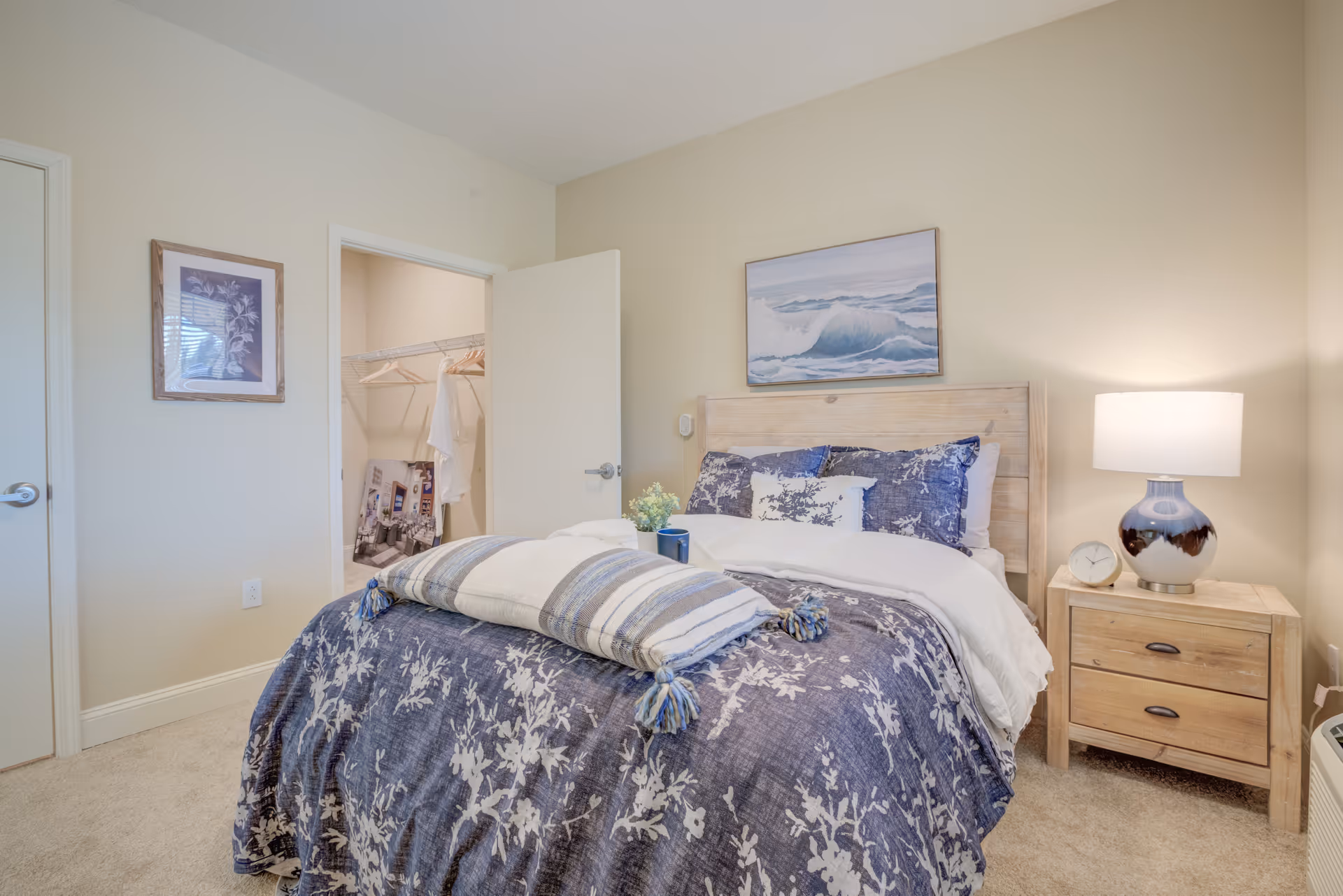 A cozy bedroom with a bed covered in blue and white floral bedding and a striped throw blanket. There is a wooden nightstand with a lamp and a small clock on it. A painting of ocean waves hangs above the bed. The room has beige walls and carpeted floor. An open door reveals a walk-in closet with hangers and some items inside.