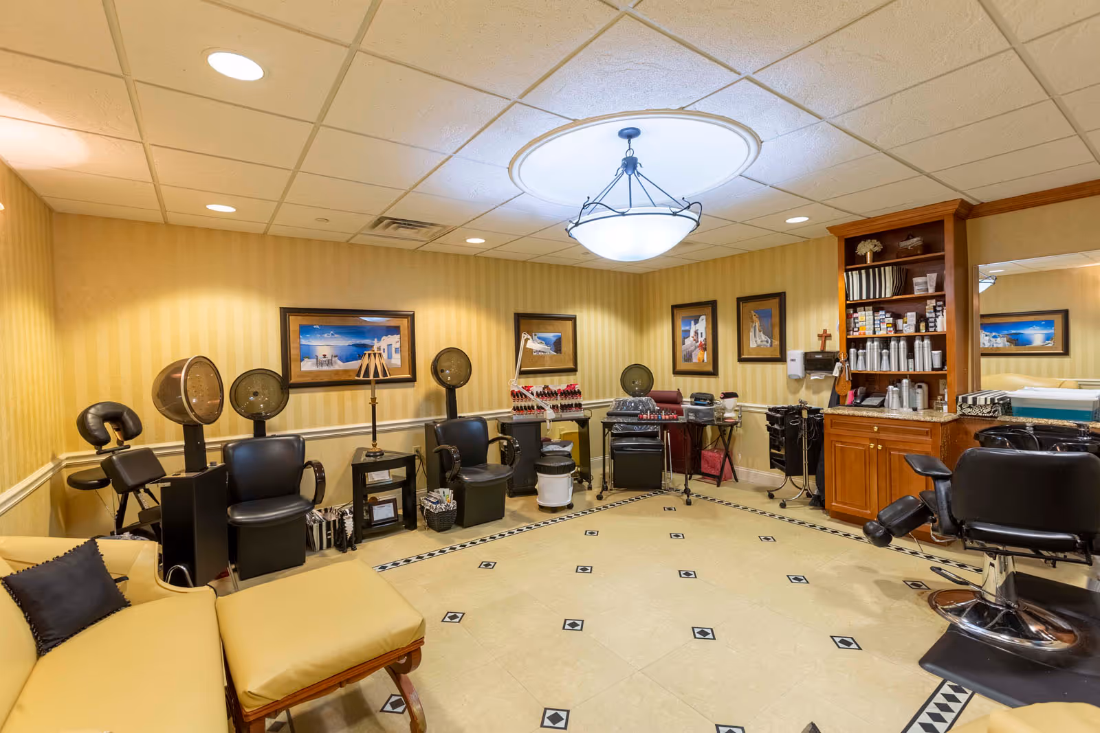 Interior view of a salon room in a senior living facility with multiple black salon chairs, hair dryers, nail polish racks, a wooden cabinet with hair products, and a yellow couch with a black pillow. The walls are yellow with framed pictures, and the ceiling has recessed lighting and a central decorative light fixture.