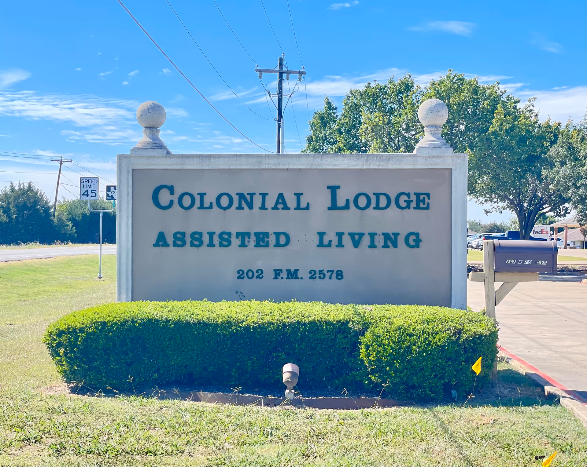 A large stone sign for Colonial Lodge Assisted Living with the address 202 F.M. 2578, surrounded by trimmed green bushes, a mailbox to the right, and a clear blue sky with some trees and a speed limit sign in the background.