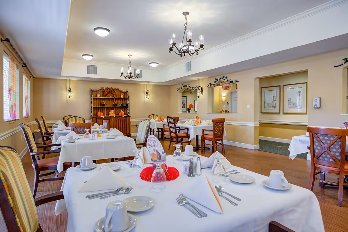 A dining room in a senior living facility with multiple tables covered in white tablecloths, set with plates, cups, utensils, and napkins. The room has wooden chairs with cushions, chandeliers hanging from the ceiling, and windows with blinds on the left side. There is a wooden hutch decorated with autumn-themed items against the far wall.