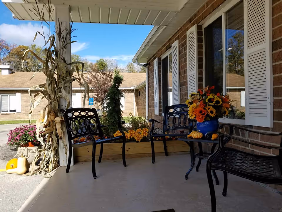 Covered front porch with black metal benches, potted flowers, pumpkins and fall decorations outside a brick building.