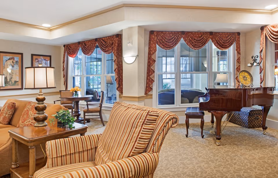 Bright communal living room with striped armchairs, a sofa, side tables, large windows, and a grand piano.