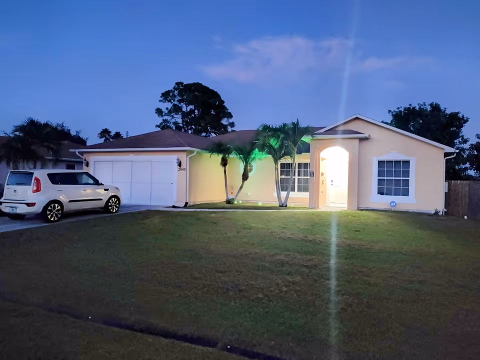 Exterior view of a single-story house at dusk with a well-maintained lawn, two palm trees near the entrance, a white car parked in the driveway, and the front door illuminated with light.