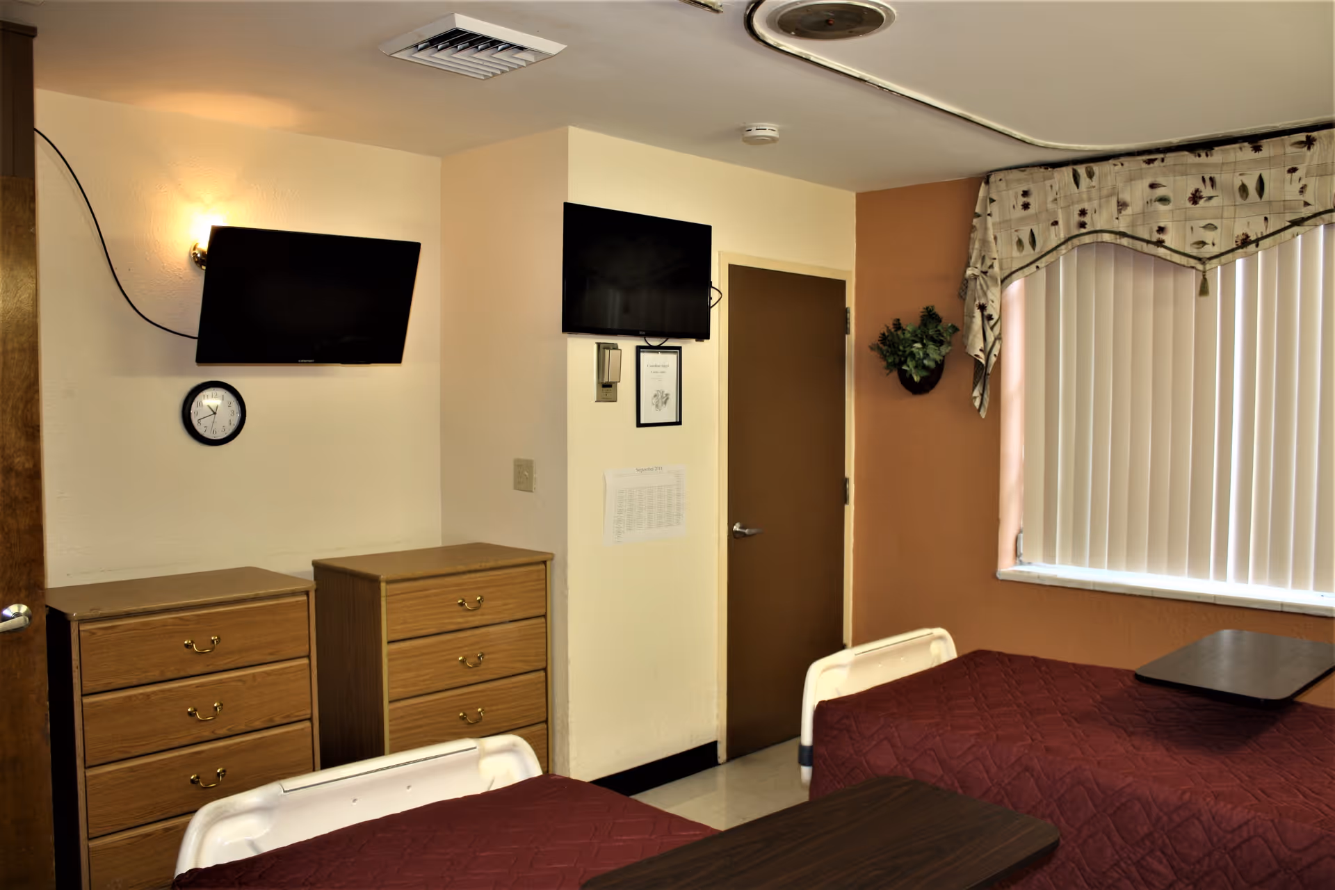 Interior view of a nursing facility room with two beds covered in maroon quilts, two wooden dressers, two wall-mounted flat-screen TVs, a clock on the wall, a door, a window with vertical blinds and a decorative valance, and a small wall-mounted plant.