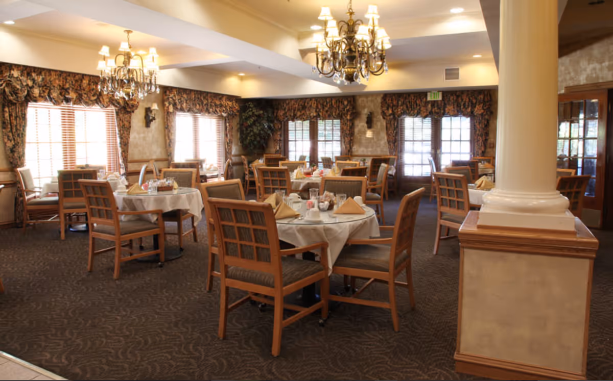 Dining room with round tables set with linens and napkins, wooden chairs, chandeliers, and curtained windows.