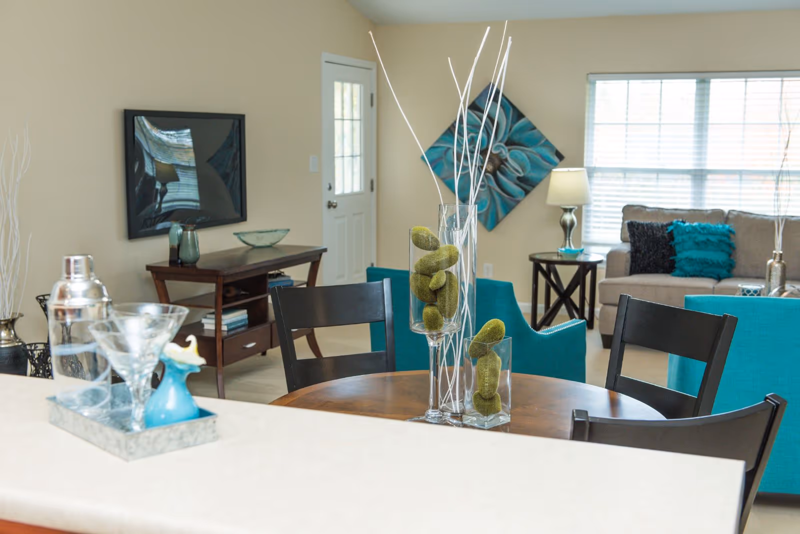 Interior view of a living room and dining area in a senior living facility. The foreground shows a wooden dining table with black chairs and decorative glass vases filled with green moss and white sticks. Behind the table, there is a beige sofa with teal and black cushions, a side table with a lamp, and a teal armchair. A wooden console table with decorative items and a wall-mounted TV are visible against a beige wall. A window with blinds allows natural light into the room.