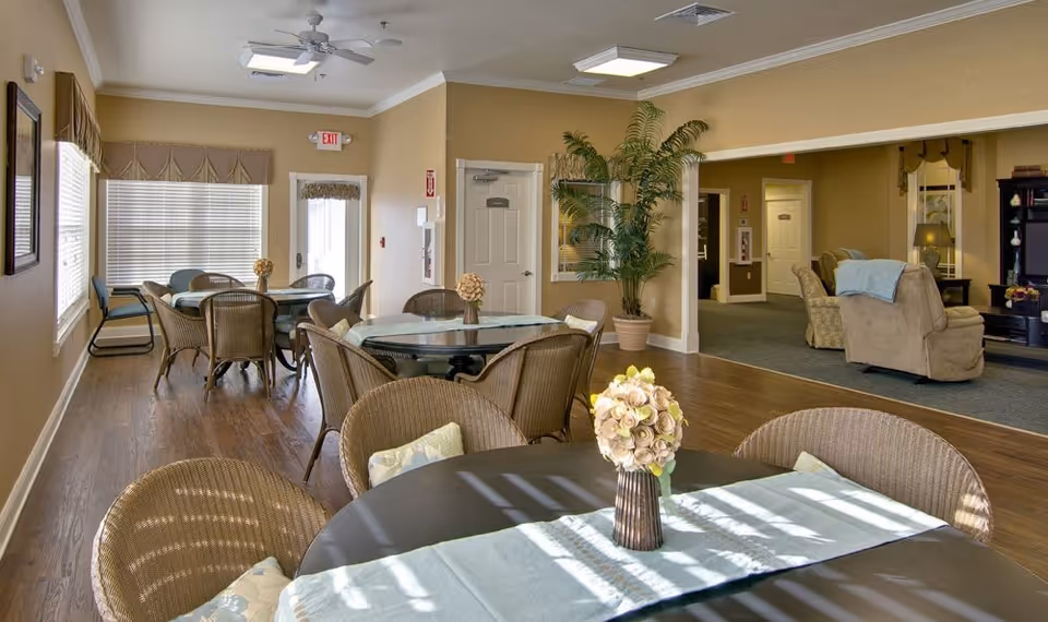 A bright and spacious common area in a senior living facility featuring multiple round tables with wicker chairs and floral centerpieces. The room has large windows with blinds and valances, wood flooring, and beige walls. In the background, there is a cozy living room area with armchairs, a lamp, and a bookshelf, separated by a wide open doorway. A large potted plant is placed near the entrance to the living room area.